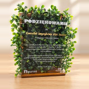 Akrylowa statuetka z podziękowaniem w drewnianej podstawie na tle zielonych listków, ze złotym napisem i tekstem